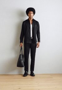 Svart langermet skjorte over en hvit tank topp, svarte snittede bukser, svarte sko og en svart tote bag, stående mot en ensfarget vegg.