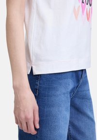 T-shirt en coton blanc avec le texte "Love You" et des graphiques de cœurs roses, avec une coupe décontractée et des fentes sur les côtés, associé à un jean en denim bleu.