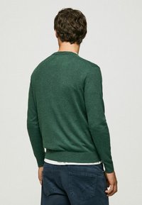 Groene gebreide trui met lange mouwen en ronde halslijn, gecombineerd met donkerblauwe broek, die een gestructureerde, gemêleerde afwerking laat zien.