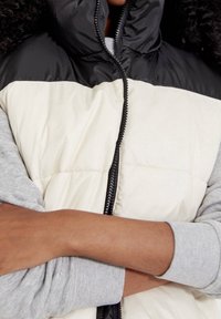 Kamizelka z ociepleniem, z czarną górną częścią i kremowym dołem, z wysokim kołnierzem, zamek błyskawiczny oraz szare mankiety. Gładka tekstura.