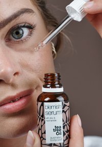 Blemish serum i en mørk amberglassflaske med en hvit pipettehett. Etiketten har rosa og svart design som fremhever tea tree olje og nøkkelingredienser.