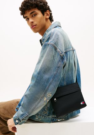 Sac bandoulière noir avec une finition texturée, doté d'une fermeture éclair et d'un accent logo. Porté sur une veste en denim à l'aspect usé.