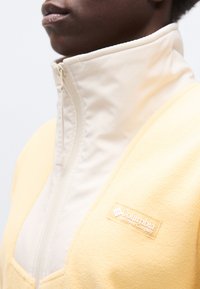 Άτομο που φοράει μια φλις ζακέτα Columbia Sportswear κίτρινη και λευκή με ψηλό γιακά που έχει κλείσει με φερμουάρ, δείχνοντας μερικώς το πρόσωπο και τον λαιμό.