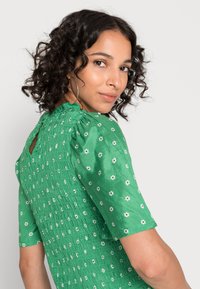 Vestido verde com um corpete franzido texturizado e mangas bufantes, apresentando um pequeno padrão floral e um detalhe nas costas em forma de olho-chave.