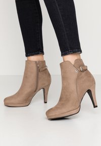Botines de ante marrón con punta afilada, tacones altos y cierre lateral de cremallera. Presenta un acento de hebilla en el lateral exterior y una textura suave.
