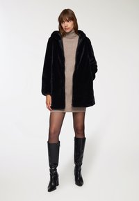 Giacca nera in pelliccia sintetica con cappuccio, chiusura a zip e taglio largo, abbinata a un vestito attillato beige e stivali neri sopra il ginocchio.