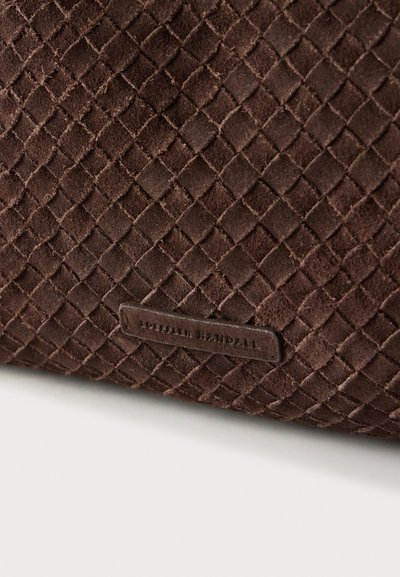 Καφέ σουέντ τσάντα με υφαντή υφή, που διαθέτει μια μικρή δερμάτινη ετικέτα με ανάγλυφο κείμενο, τονίζοντας το μοναδικό της σχέδιο και την τεχνική της κατασκευή.