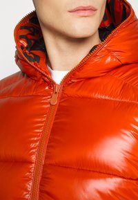 Leuchtend orangefarbene gepolsterte Jacke mit einem glänzenden Finish, ausgestattet mit einem hohen Kragen, Reißverschluss und einem gemusterten Innenfutter.