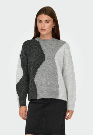 Vrouw in een grijs sweater met lange mouwen en kleurblokken, gecombineerd met een zwarte rok, staande tegen een effen lichte achtergrond.