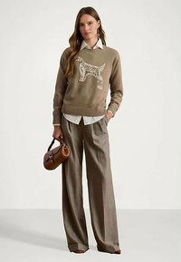 Γυναίκα φορώντας ένα μπεζ πουλόβερ με σχέδιο σκύλου, λευκό πουκάμισο, παντελόνι φαρδύ, κρατώντας μια καπνίτσα τσάντα, στέκεται μπροστά σε ένα απλό φόντο.