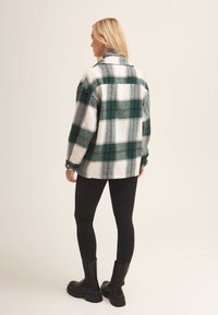 Green and white plaid flannel shirt with long sleeves, button cuffs, and a rounded hem, paired with black leggings and chunky black boots.