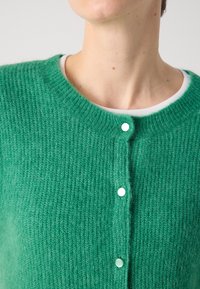 Gros plan d'une personne portant un cardigan tricoté vert avec trois boutons fermés sur un t-shirt blanc à col rond, sur un fond neutre.