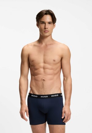 Junger Mann mit trainiertem Körper trägt marineblaue Hugo-Boxershorts und steht vor einem einfarbigen weißen Hintergrund.