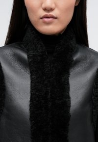 Μαύρο εξωτερικό ένδυμα από τεχνητό δέρμα με μαλακό, υφασμένο μαύρο φλις γιακά; διαθέτει ψηλή λαιμόκοψη και συμμετρικό σχέδιο.