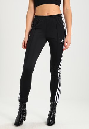 Kobieta w czarnych spodniach dresowych Adidas z białymi paskami po bokach i czarnych botkach na obcasie, stojąca na jednolitym tle.