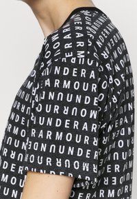 Camiseta negra con un patrón de texto blanco en toda la prenda que presenta la palabra "UNDER ARMOUR" en diferentes orientaciones y tamaños. Mangas cortas, cuello redondo.