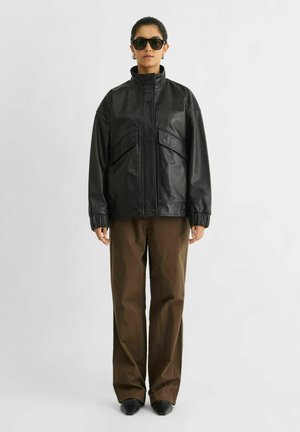 Un mannequin se tient devant un fond clair, vêtu d’une veste en cuir noire, d’un pantalon marron, de chaussures noires et de lunettes de soleil foncées.