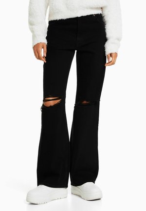 Personne portant un jean évasé noir avec des genoux déchirés, un pull blanc duveteux et des baskets blanches compensées, debout devant un fond uni.