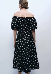 Rochie neagră cu umerii descoperiți, cu buline albe, având mâneci bufante, talie strânsă și fustă de lungime medie.
