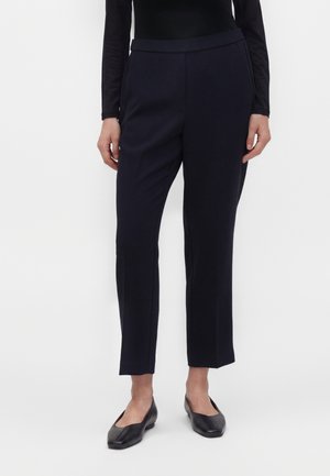Femme portant un pantalon noir ajusté aux chevilles, un haut noir à manches longues et des chaussures plates noires, debout devant un fond uni.