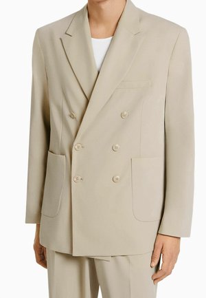 Uomo che indossa un blazer doppiopetto beige con sei bottoni, rever a lancia, tasche applicate e pantaloni beige abbinati sopra una camicia bianca.