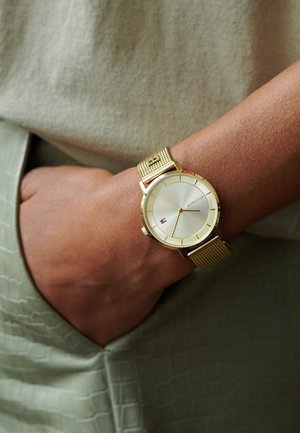 Gold mesh wristwatch with a white face, featuring a minimalist design and subtle hour markers. The watch displays a small logo at 6 o'clock.