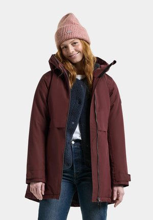 Smilende kvinde med langt rødt hår iført en lyserød strikhue, bordeaux vinterjakke, marineblå fleecejakke, hvid skjorte og blå jeans.