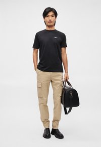 Melnas īsām piedurknēm T-krekls ar logotipu, bēšas kravas bikses ar sānu kabatām un melna soma ar brūnām ādas akcentiem. Melni potīšu zābaki.