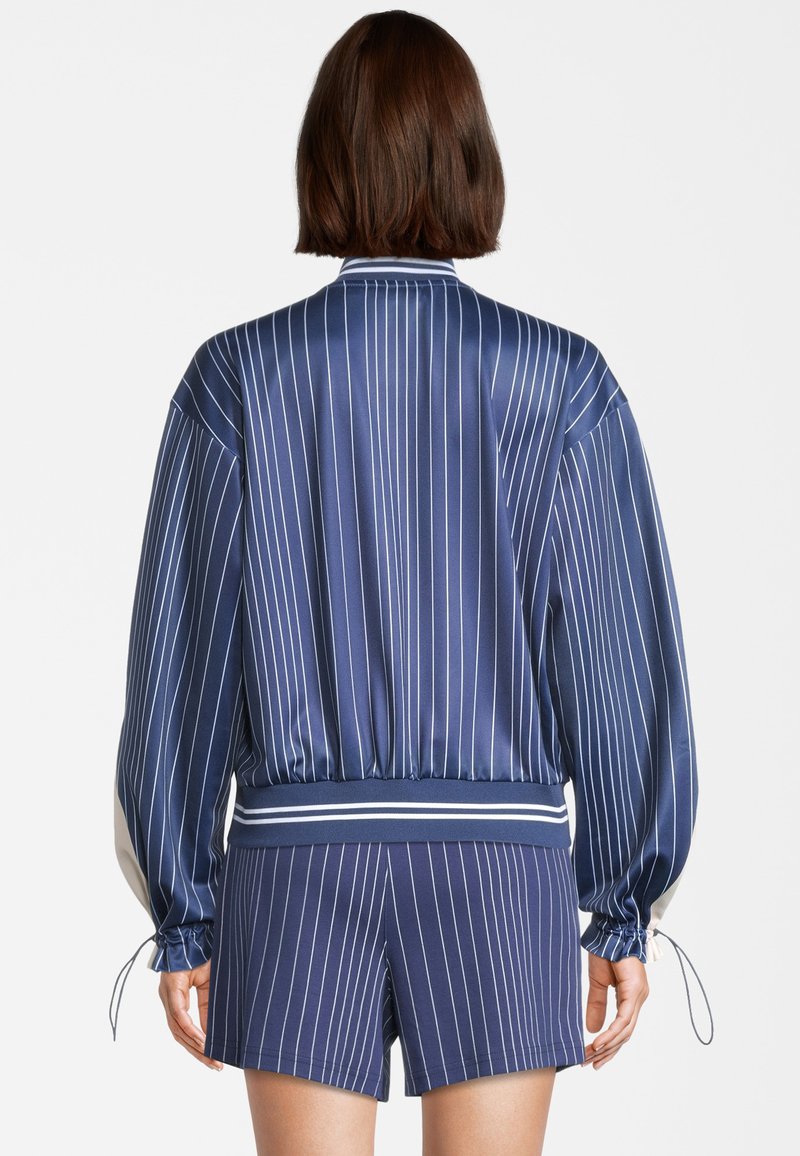 Giacca bomber in satin blu navy con strisce verticali bianche, vita elasticizzata e maniche arricciate, abbinata a pantaloni shorts coordinati.