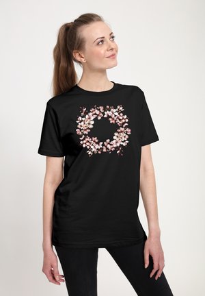 Femme debout, regardant vers le haut, portant un t-shirt noir avec un motif floral circulaire rose et blanc sur le devant, fond uni.