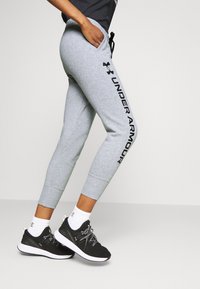 Joggers deportivos grises con un ajuste ajustado, que presentan un logo negro en el lateral. Combinados con zapatillas negras y calcetines blancos hasta el tobillo.