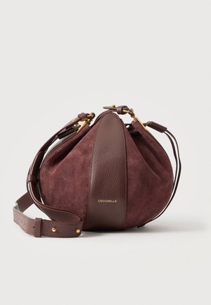 Sac à main rond en daim et cuir brun foncé avec bandoulière réglable et ferrures dorées, marqué « Coccinelle » sur le panneau avant.