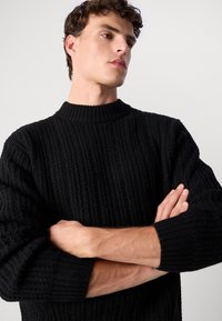 Young man with light skin and brown hair wearing a black textured knit sweater, arms crossed, looking to the side against a plain background.