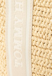 Geflochtener Strohtasche in hellbeiger Farbe, mit strukturierter Oberfläche und einem weißen Stoffriemen mit geprägter Schrift.