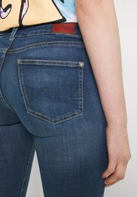 Los jeans de mezclilla azul oscuro presentan un diseño clásico de cinco bolsillos, con una forma distintiva en el bolsillo trasero y sutiles acentos de costura.
