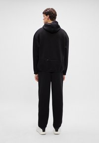 Muž oblečený v čiernej mikine a čiernych nohaviciach, otočený chrbtom, stojí na bielom podlahe s jednoduchým bielym pozadím.