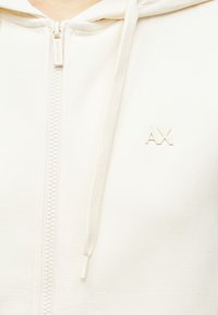 Šviesiai kreminė megztinė su priekiu užtrauktuku, su mažuoju baltu "AX" logotipu ir su raišteliais, pagaminta iš minkštos medvilnės medžiagos.