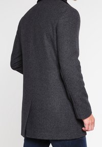 Uomo che indossa un cappotto di lana grigia scura e testurizzata con una fessura posteriore, in piedi contro uno sfondo bianco, mano parzialmente visibile.