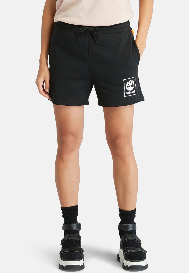 Shorts en coton noir avec une taille élastique et un cordon de serrage, arborant un logo Timberland rectangulaire blanc. Texture lisse, longueur mi-cuisse.