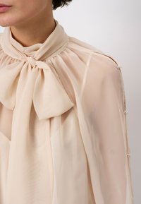 Helle beige Bluse aus transparentem Stoff. Sie hat einen hohen Kragen mit einer Schleifendetail und Knopf-Akzenten entlang der Schultern. Weicher, fließender Stoff.
