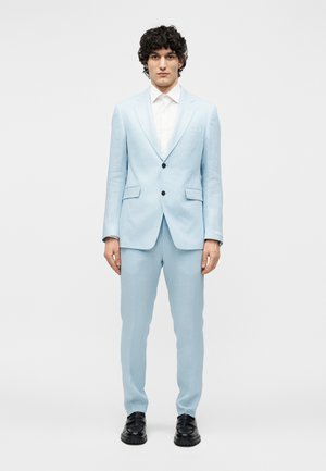 Mand med krøllet hår iført en lyseblå jakkesæt, hvid skjorte og sorte sko, stående mod en ensartet lys baggrund.