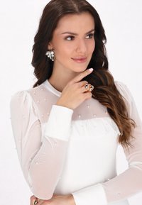 Top bianco con maniche lunghe trasparenti decorate con perle, un design frontale arruffato e un corpo in tessuto liscio e solido.