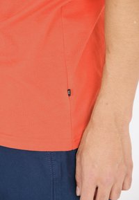 Kortärmad, korallfärgad t-shirt i bomull. Har en stickad fåll och en liten svart etikett på sidan. Bärs med mörkblå shorts.