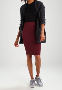 Mujer vistiendo una blusa negra, un cárdigan de punto gris oscuro, una falda lápiz burdeos y zapatillas plateadas, de pie contra un fondo liso.