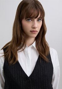 Czarna kamizelka w drobną prążka, noszona na białej koszuli zapinanej na guziki, z dekoltem w serek i pojedynczym zapięciem na guzik. Gładka tkanina o teksturowanym wykończeniu.