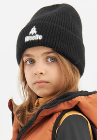 Svart ribbad beanie med vit "WeeDo"-logga; bärs med en jacka i orange och svart. Har mellanlånga bruna hår som ramar in ansiktet.