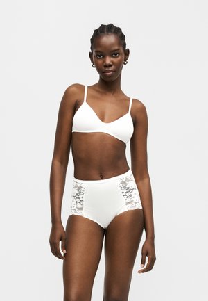 Mujer con el pelo trenzado que lleva un bralette blanco y ropa interior blanca de talle alto con paneles de encaje, de pie contra un fondo claro y liso.