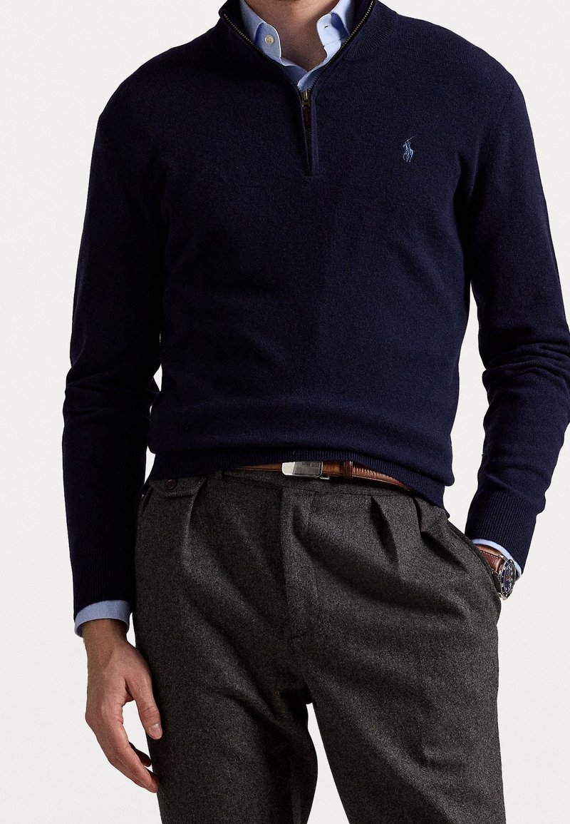 Homme portant un pull bleu marine à fermeture éclair jusqu'au milieu du cou par-dessus une chemise à col bleu clair, un pantalon gris plissé, une ceinture marron et une montre au poignet gauche.