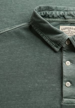 Polo verde con colletto piegato e tre bottoni bianchi, che mostra parte dell'etichetta del marchio Superdry all'interno del colletto.