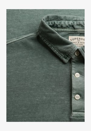 Polo verde con colletto piegato e tre bottoni bianchi, che mostra parte dell'etichetta del marchio Superdry all'interno del colletto.